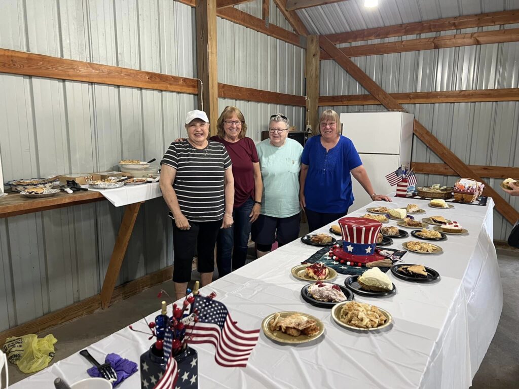 four women standing behind a table of food decorated for July 4th